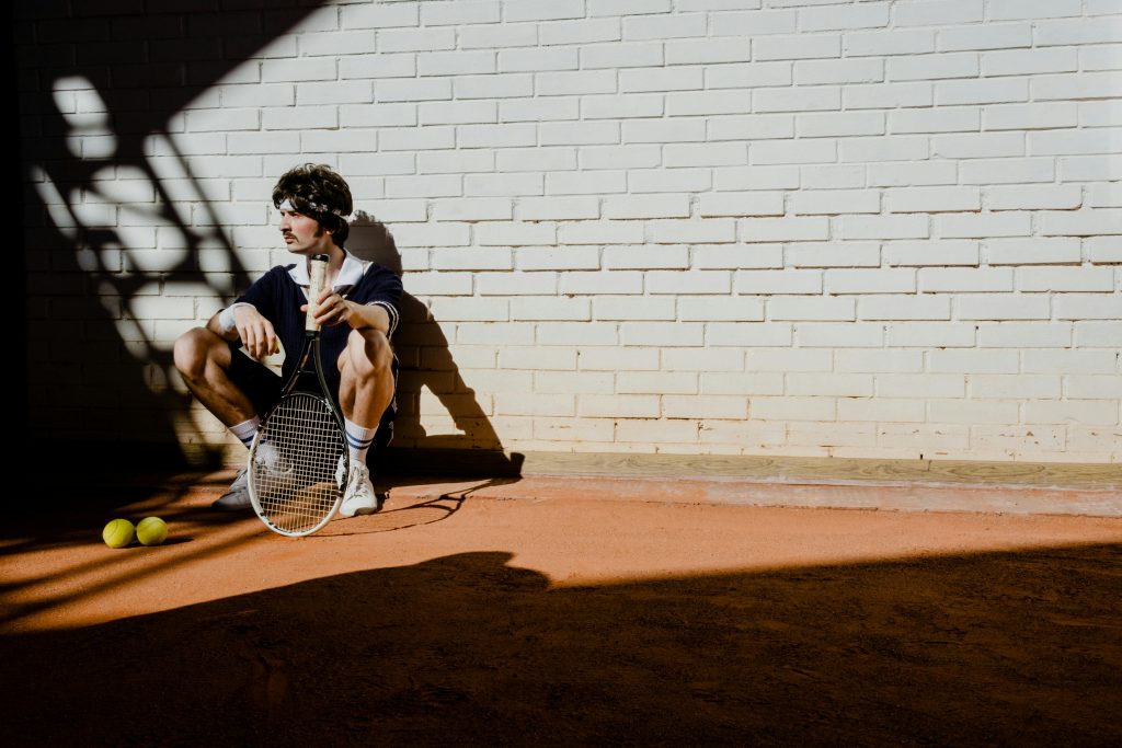 A stylish man in vintage sportswear sits with a tennis racket and balls, casting shadows.
